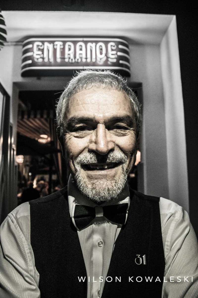 Black and white portrait of a grinning silver haired doorman under a neon Entrance sign